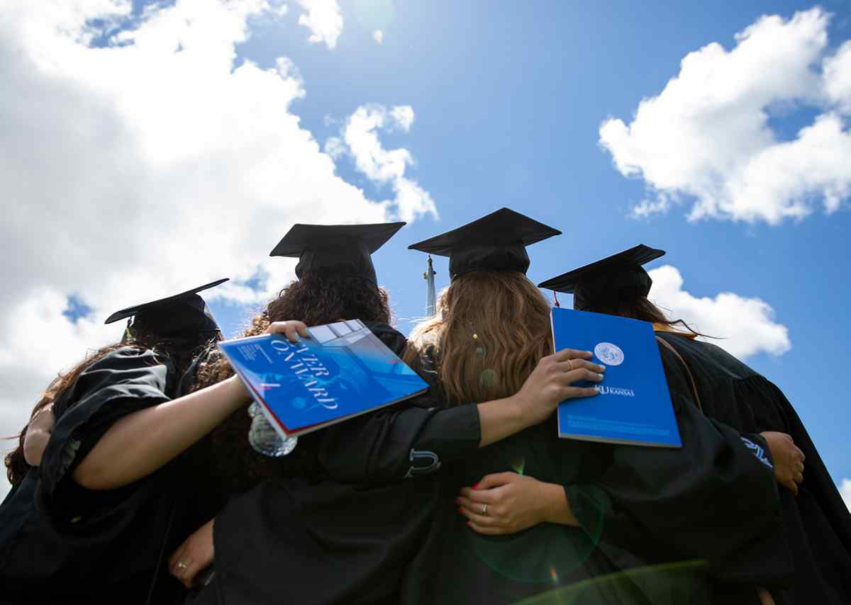 Four KU graduates with arms around one another. The photo is shot from behind showing the student's backs and embracing arms, two student are holding commencement programs.