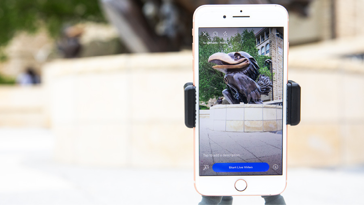 Iphone in a tripod on campus shows Facebook Live interface with a Jayhawk statue in view